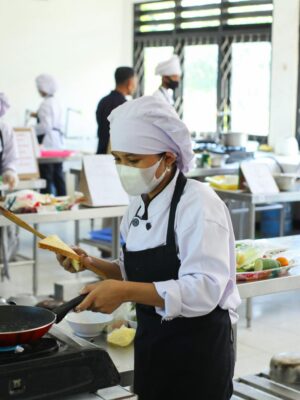 Chefs practicing culinary skills in a training kitchen setting, wearing masks for safety.